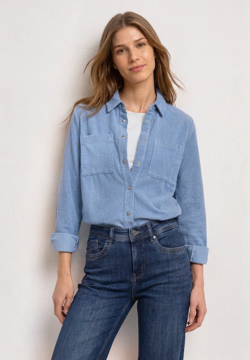 Young woman with light brown hair wearing a light blue corduroy shirt over a white top and dark blue jeans standing against a white wall.
