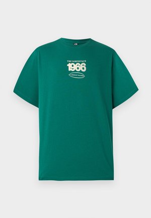 Person mit dunklem lockigem Haar und Vitiligo an den Armen, trägt ein türkisfarbenes The North Face T-Shirt mit der Aufschrift "1966" und beige Cargohosen, von hinten gesehen.