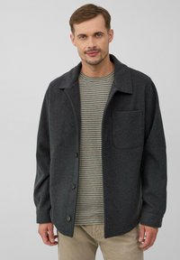 Donkergrijze button-up jas van gestructureerde stof, met een kraag en een enkele borstzak, gedragen over een gestreept shirt.