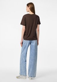 Femme debout de dos, portant un t-shirt marron foncé à manches courtes, un jean large bleu clair et des baskets argentées sur fond blanc.