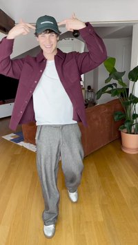 Cappellino verde, maglietta color granata, t-shirt bianca, pantaloni grigi con motivo, e scarpe da ginnastica argento. Ambiente interno con piante e un divano. Abbigliamento casual.