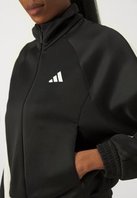 Giacca nera con zip e colletto alto, dotata di dettagli in logo bianco e polsini elasticizzati. Tessuto liscio e leggermente lucido con tasche laterali.