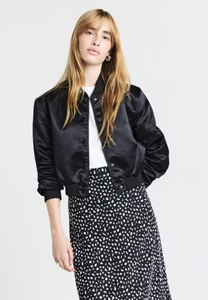 Young woman with long blonde hair wearing black satin bomber jacket, white shirt, and black skirt with white abstract spots, posing against white background.