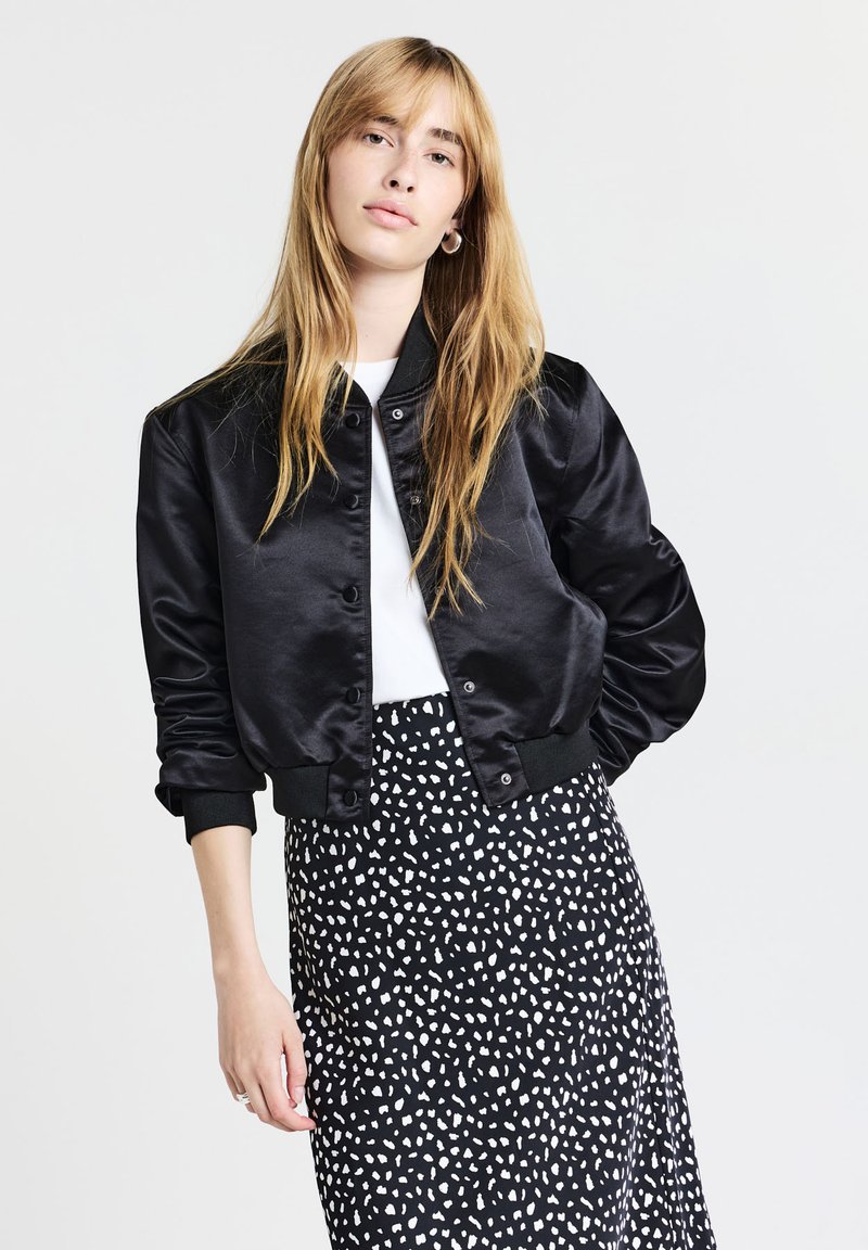 Jeune femme aux longs cheveux blonds portant une veste bomber en satin noir, une chemise blanche et une jupe noire avec des taches abstraites blanches, posant devant un fond blanc.