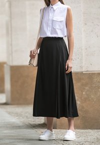 Donna che indossa una camicia senza maniche bianca con colletto, una gonna midi nera, sneaker bianche e una borsa a spalla beige, in piedi all'aperto.