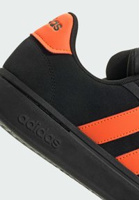 Sneaker nera con accenti arancioni, caratterizzata da un upper sintetico, tre strisce texturizzate e suola in gomma con logo del marchio in rilievo.