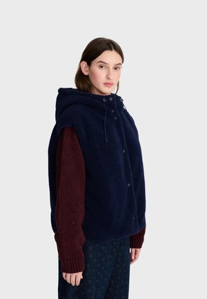Mujer con un chaleco de forro polar azul marino con capucha sobre un suéter burdeos y pantalones oscuros con estampado, de pie frente a un fondo claro y liso.