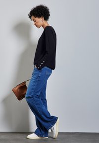 Maglione nero con dettagli a bottone sulle maniche, abbinato a jeans a zampa d'elefante blu e sneakers bianche. La modella tiene una borsa marrone, in piedi di lato.