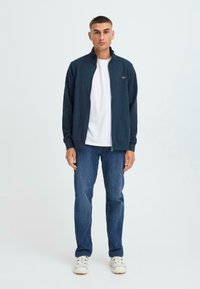 Jūras spēku zip-up jaka ar augstu apkakli, kombinēta ar baltu t-kreklu un zilām džinsām. Jaka rotāta ar mazu logo uz krūtīm.