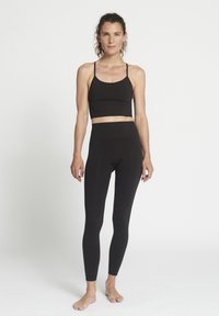 Donna in piedi a piedi nudi, con un crop top nero aderente e leggings a vita alta, contro uno sfondo bianco.