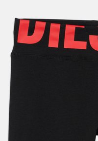 Juodos tamprės su storu raudonu logotipo juostos diržu ties liemene, pasižyminčios lygiu paviršiumi ir elastinga medžiaga, suteikiančia komfortą ir lankstumą.