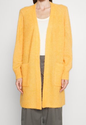 Vrouw draagt een lange, zachte gele cardigan met voorzakken, wit topje en grijze broek, staand tegen een effen achtergrond.