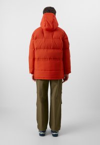 Oransje pufferjakke med hette, med quiltet tekstur og avslappet passform. Kombinert med løse olivengrønne cargo-bukser og blå joggesko.
