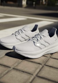 Coppia di scarpe da corsa Adidas bianche con suole Boost posizionate su un marciapiede piastrellato vicino a un attraversamento pedonale.
