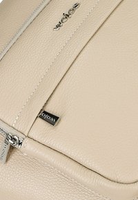 Beige leren rugzak met een gestructureerd oppervlak, zilveren hardware en een ritsluiting. Voorzien van subtiele stiksels en een merklabel.