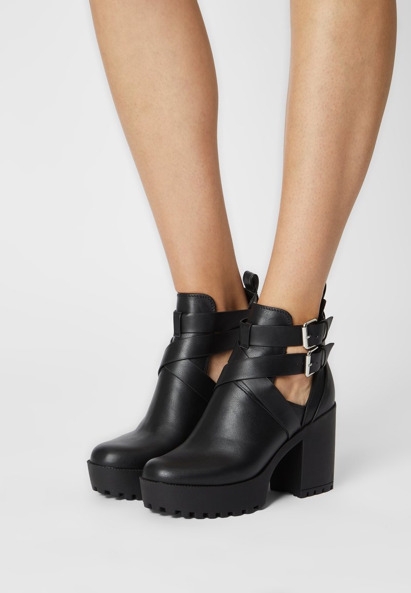 Botas negras hasta el tobillo hechas de cuero con gruesos tacones en bloque, acentos de doble hebilla y correas cruzadas. Suela texturizada para mayor tracción.