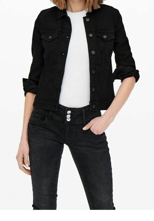 Frau trägt eine taillierte schwarze Jeansjacke mit hochgekrempelten Ärmeln, weißes T-Shirt und schwarze Jeans mit dreifacher Knopfleiste, Hand in der Tasche.