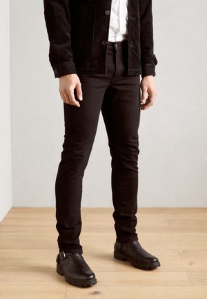 Black corduroy jacket over a white shirt, paired with slim-fit black pants and chunky black boots. Wood floor background.