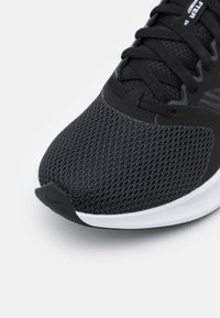 Zapatilla deportiva negra con parte superior de malla, tejido texturizado y suela blanca. Presenta un cordón tradicional y una puntera reforzada.