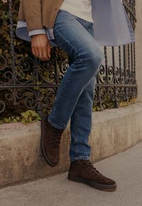 Személy barna velúr magas szárú sneakerben és kék farmerruhában, az egyik lábát egy kőkorlátnak támasztva, háttérben díszes fémkerítéssel.