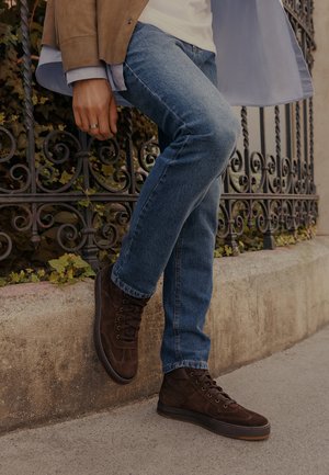 Persona che indossa sneaker alte in camoscio marrone e jeans blu, appoggiando una gamba contro un muretto di pietra con uno sfondo di una recinzione metallica ornamentale.