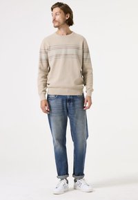 Beige Strickpullover mit horizontalen blauen und grauen Streifen, kombiniert mit hellblauen Jeans und weißen Sneakers, mit umgeschlagenen Manschetten.
