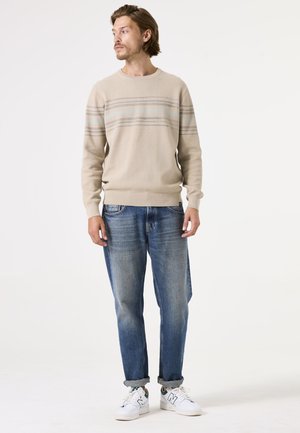 Suéter de punto beige con rayas horizontales en azul y gris, combinado con jeans de mezclilla azul claro y zapatillas blancas, con puños enrollados.