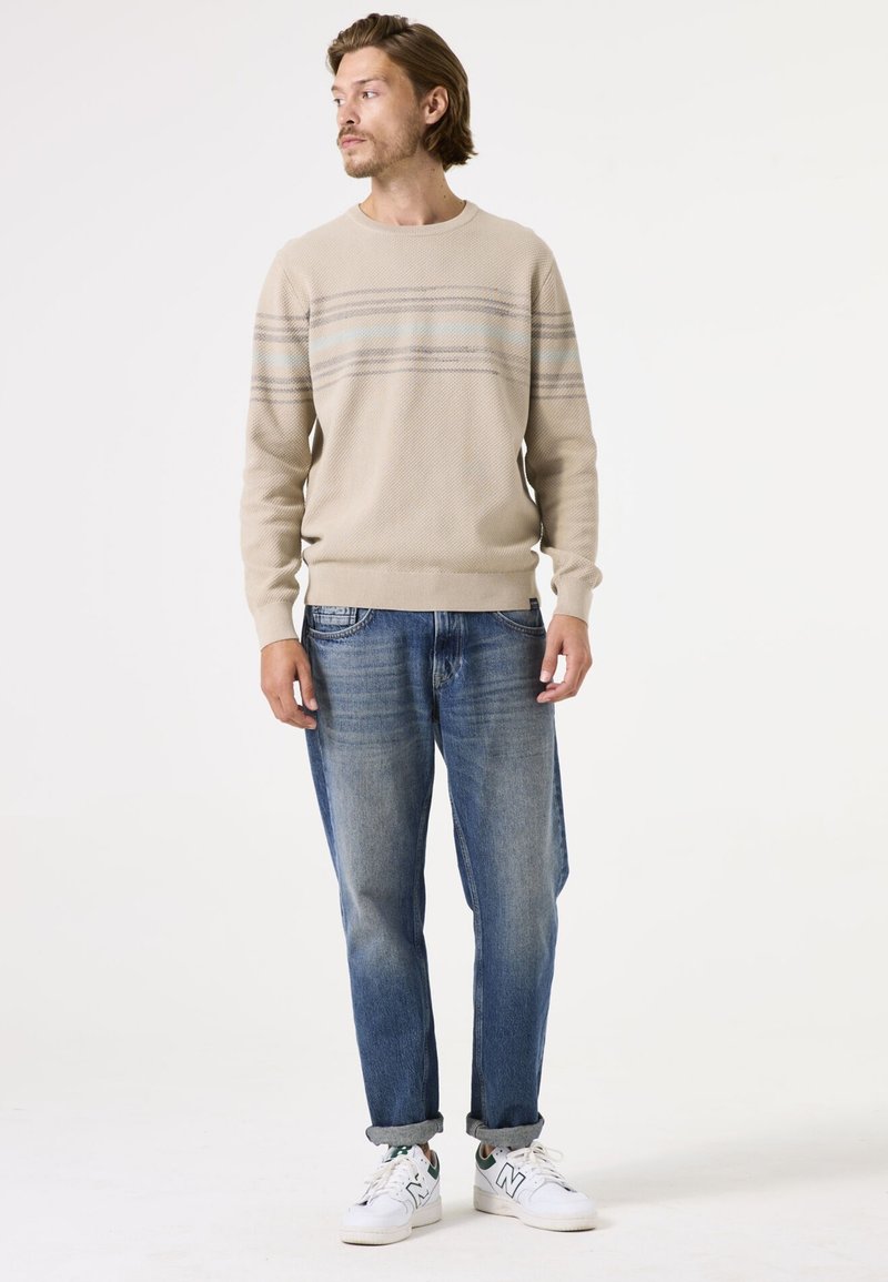 Beige Strickpullover mit horizontalen blauen und grauen Streifen, kombiniert mit hellblauen Jeans und weißen Sneakers, mit umgeschlagenen Manschetten.