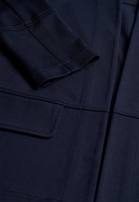 Tummansininen kangas, jossa näkyvät ompeleet ja taitettu hihansuu suorakulmaisen taskuläppä vieressä vaatteessa.