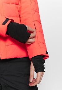 Main ajustant la fermeture éclair de la manche d'une veste matelassée orange vif, portant des gants noirs sans doigts et un pantalon noir.