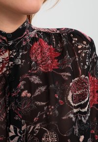Blusa nera con scollatura a bottoni che presenta motivi floreali rossi e rosa. Realizzata in tessuto leggero e trasparente con una sottile texture.