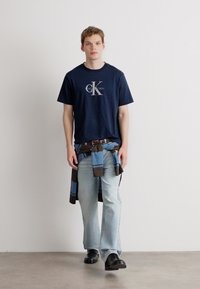 Námořnické bavlněné tričko s logem, světle modré džíny a modro-hnědá kostkovaná flanelová košile uvázaná kolem pasu. Černé boty doplňují outfit.