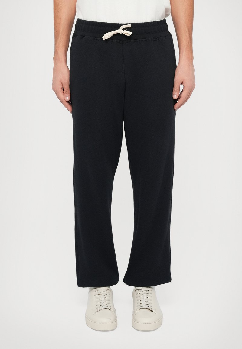 Schwarze Jogginghosen mit weicher Textur, elastischem Bund und verstellbarem Kordelzug. Kombiniert mit hellen Sneakers.
