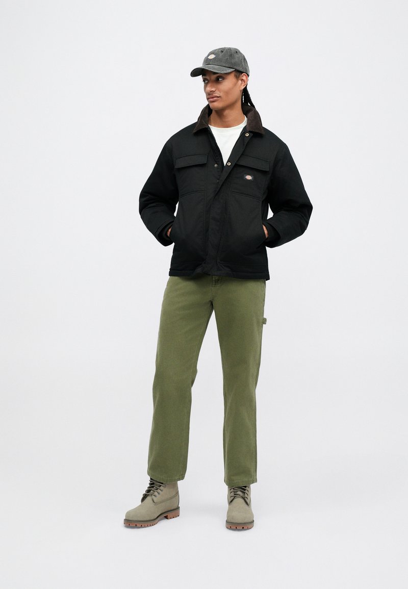 Uomo con giacca nera, camicia bianca, pantaloni verde oliva, stivali beige e berretto grigio scuro, in piedi con le mani nelle tasche della giacca, che guarda a destra.