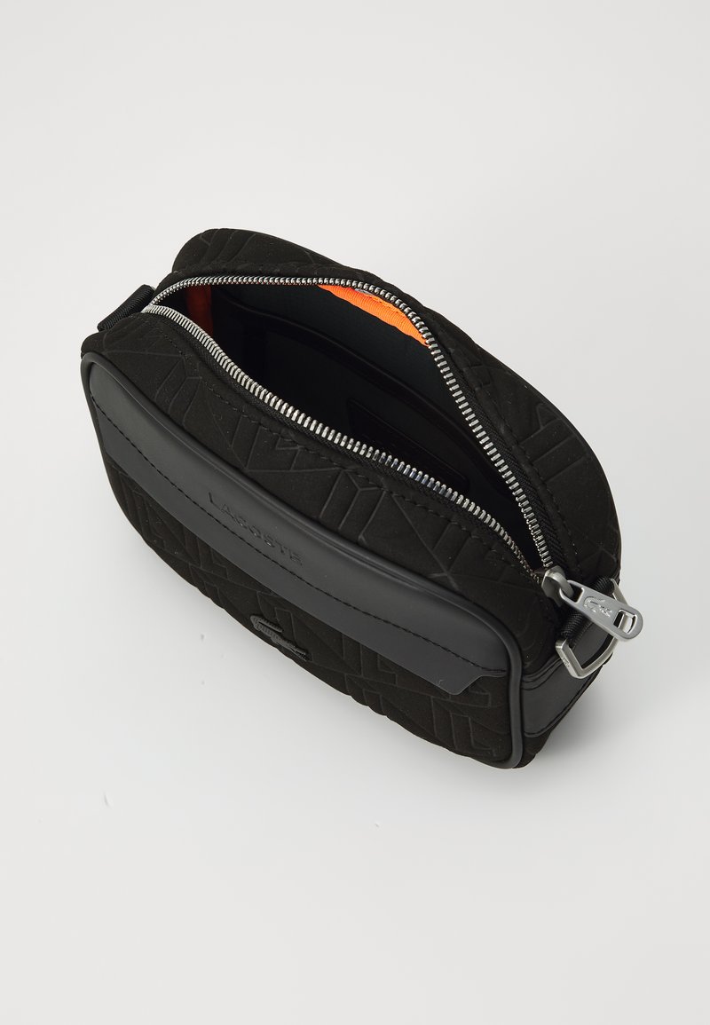 Sac en tissu noir texturé avec fermeture éclair, intérieur orange et accent en cuir noir lisse. Présente un logo en relief.