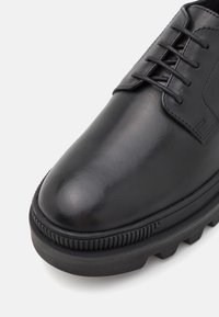 Chaussure habillée en cuir noir présentant une finition lisse, un bout rond et une semelle en caoutchouc texturé avec un motif de relief prononcé.