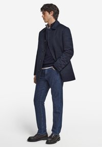 Marineblauer Wollmischmantel mit Knopfleiste, darüber ein marineblauer Pullover und ein helles Hemd, kombiniert mit dunkelblauer Jeans und schwarzen Schuhen.