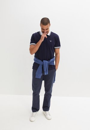 Polo shirt blu navy con accenti bianchi, indossato con un pullover azzurro chiaro legato in vita, abbinato a pantaloni scuri e sneakers bianche.