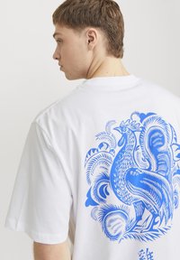 Camiseta de algodón blanca con un gran gráfico azul de un gallo estilizado en la parte posterior, con patrones y texturas intrincadas.