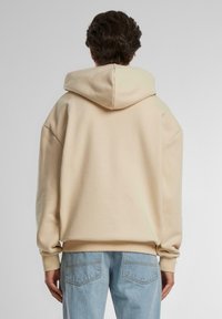 Beige hooded sweatshirt with a relaxed fit, featuring dropped shoulders and ribbed cuffs. Soft texture, simple design, and no visible patterns.