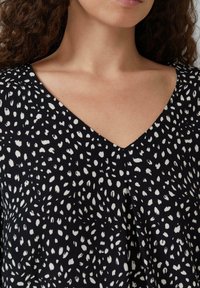 Blouse noire avec un col en V, fabriquée en tissu léger, ornée d'un motif à pois abstraits blancs, mettant en valeur des plis à l'avant.