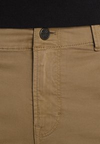 Gros plan sur un pantalon kaki à l'avant avec un bouton noir et des coutures visibles, porté avec une chemise noire côtelée rentrée.