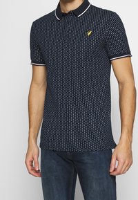 Navy polo shirt with white polka dots, short sleeves, and a contrasting collar. Features a small yellow logo on the chest.