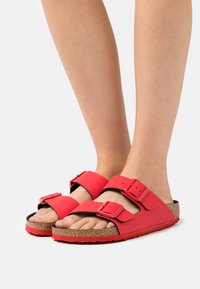 Punainen kaksihihnainen liukukenkä, jossa on säädettävät soljet, korkkinen pohja ja teksturoitu kumipohja. Sileä pinta ja avonainen kärki.