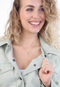 Gouden ketting met een heldere, ronde hanger, gedragen over een witte top en een lichtgroene jas met een kraag en grijze knopen. Getextureerde stof.