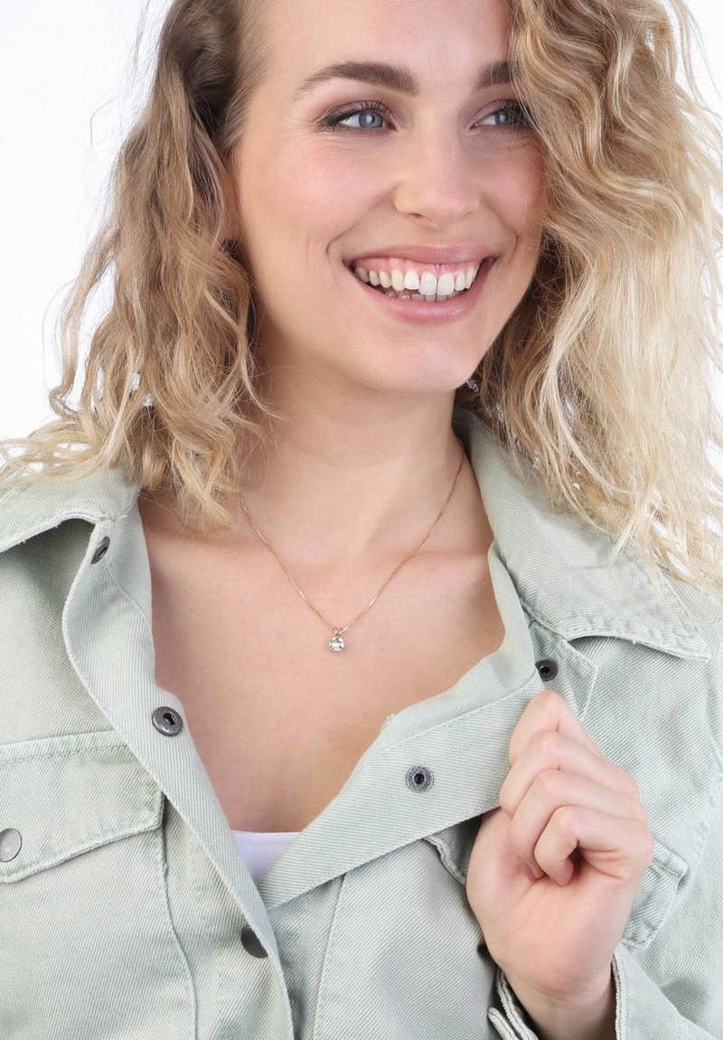 Gouden ketting met een heldere, ronde hanger, gedragen over een witte top en een lichtgroene jas met een kraag en grijze knopen. Getextureerde stof.