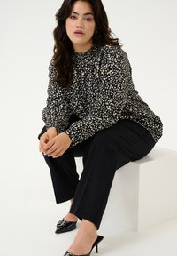 Juoda bluzė su balta ir auksine gėlių raštu, sudeginta apykaklė, ilgomis rankovėmis, derinama su juodos spalvos kelnėmis ir smailiomis aukštakulnėmis bateliais.