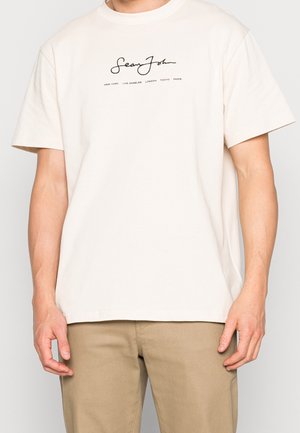 T-shirt en coton de couleur crème avec un col rond, des manches courtes et un texte en logo noir sur le devant qui indique "Sean John" et des noms de villes.