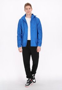 Giacca blu con un motivo geometrico, materiale leggero, dotata di cappuccio e cordoncini regolabili; abbinata a pantaloni neri e sneakers.