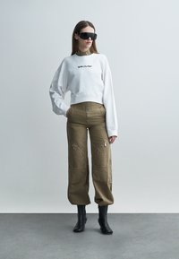 Femeie purtând un hanorac alb supradimensionat, pantaloni cargo kaki cu detalii de fermoar, cizme negre cu toc și ochelari de soare negri mari, stând pe un podea gri.